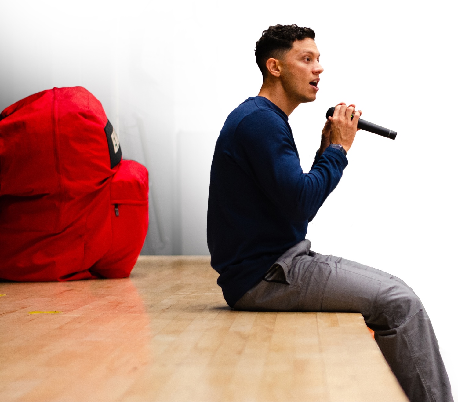 Education keynote speaker Sam Demma sitting on the edge of a stage talking into a microphone with his big red backpack just behind him on stage